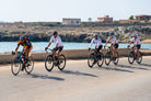 Qunafa team cyclists riding on a road near a waterfront Mediterranean sea on the way to Syracuse, Sicily Italia. Tour Guided Experience by Qunafa Travel